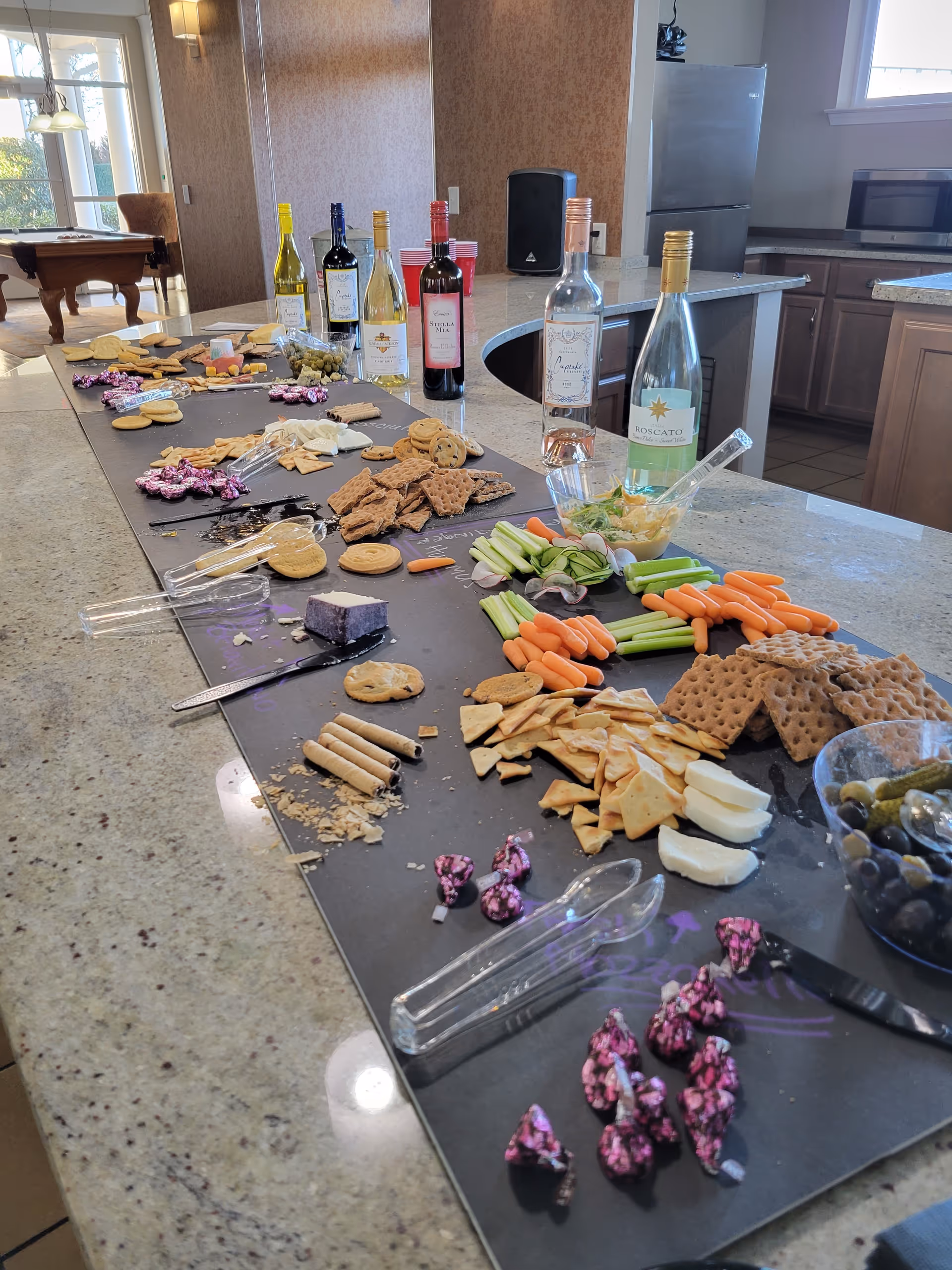 A kitchen island laden with cheeses, crackers, veggies, and multiple wine bottles.