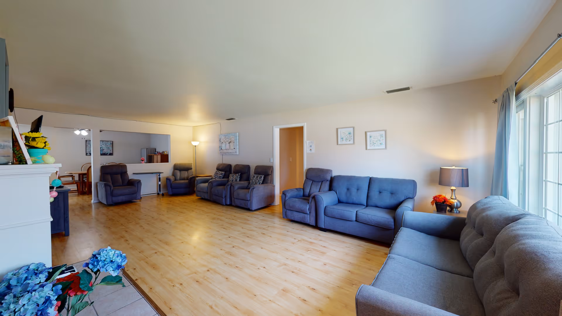 A spacious living room in an elderly care facility with multiple gray upholstered sofas and recliners arranged along the walls. The room has light wood flooring, beige walls, and a large window with curtains letting in natural light. There is a floor lamp, framed artwork on the walls, and a small table with a lamp and flowers near the window. In the background, a dining area with a table and chairs is partially visible through a large interior window opening.