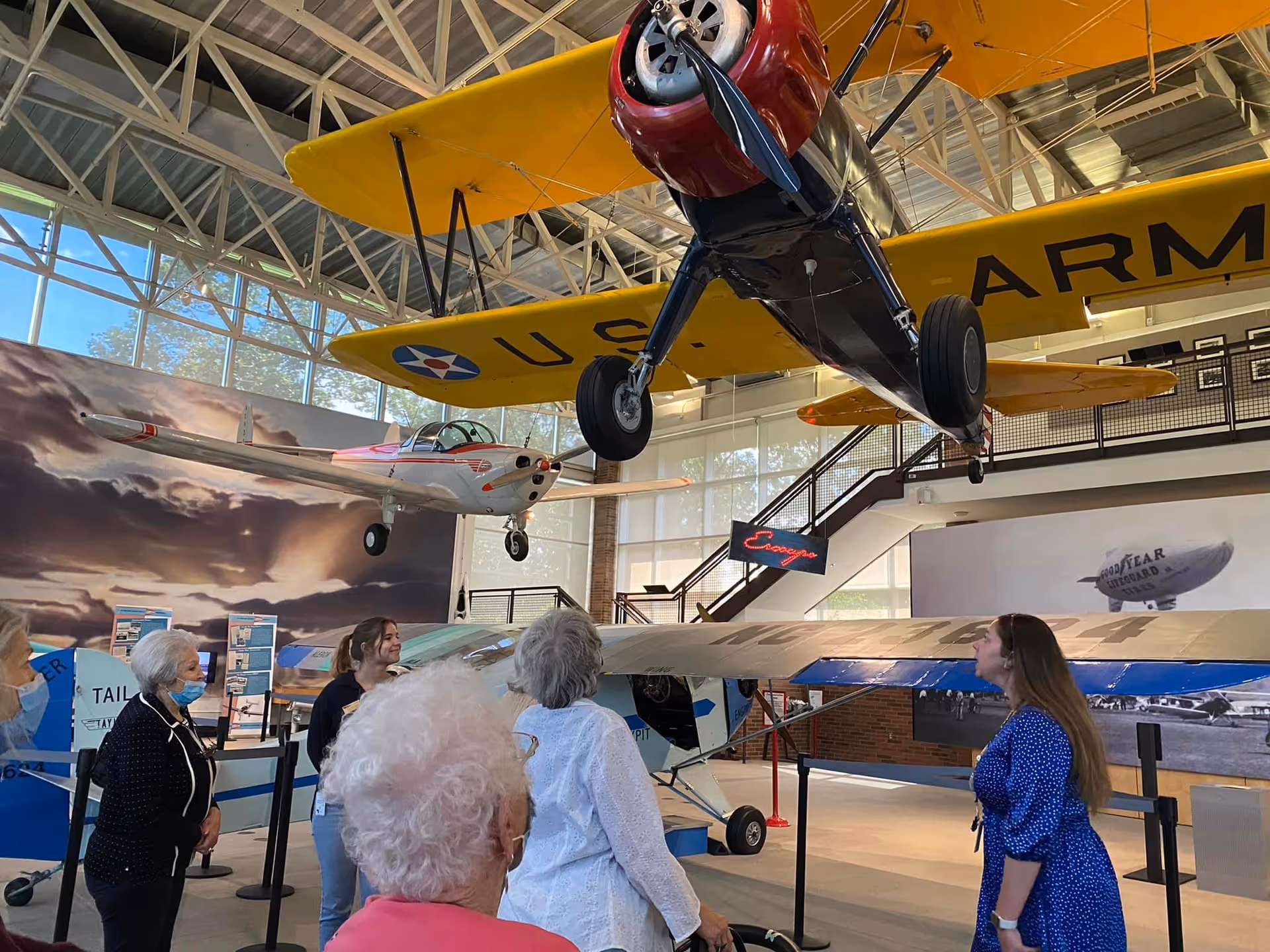 A group of elderly people and a younger woman are inside an aviation museum looking at vintage airplanes suspended from the ceiling and displayed on the floor. The museum has large windows and a high ceiling with metal beams.