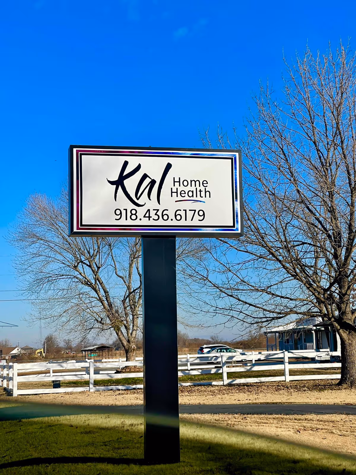 Outdoor Kal Home Health sign on a tall post with trees and a white fence.