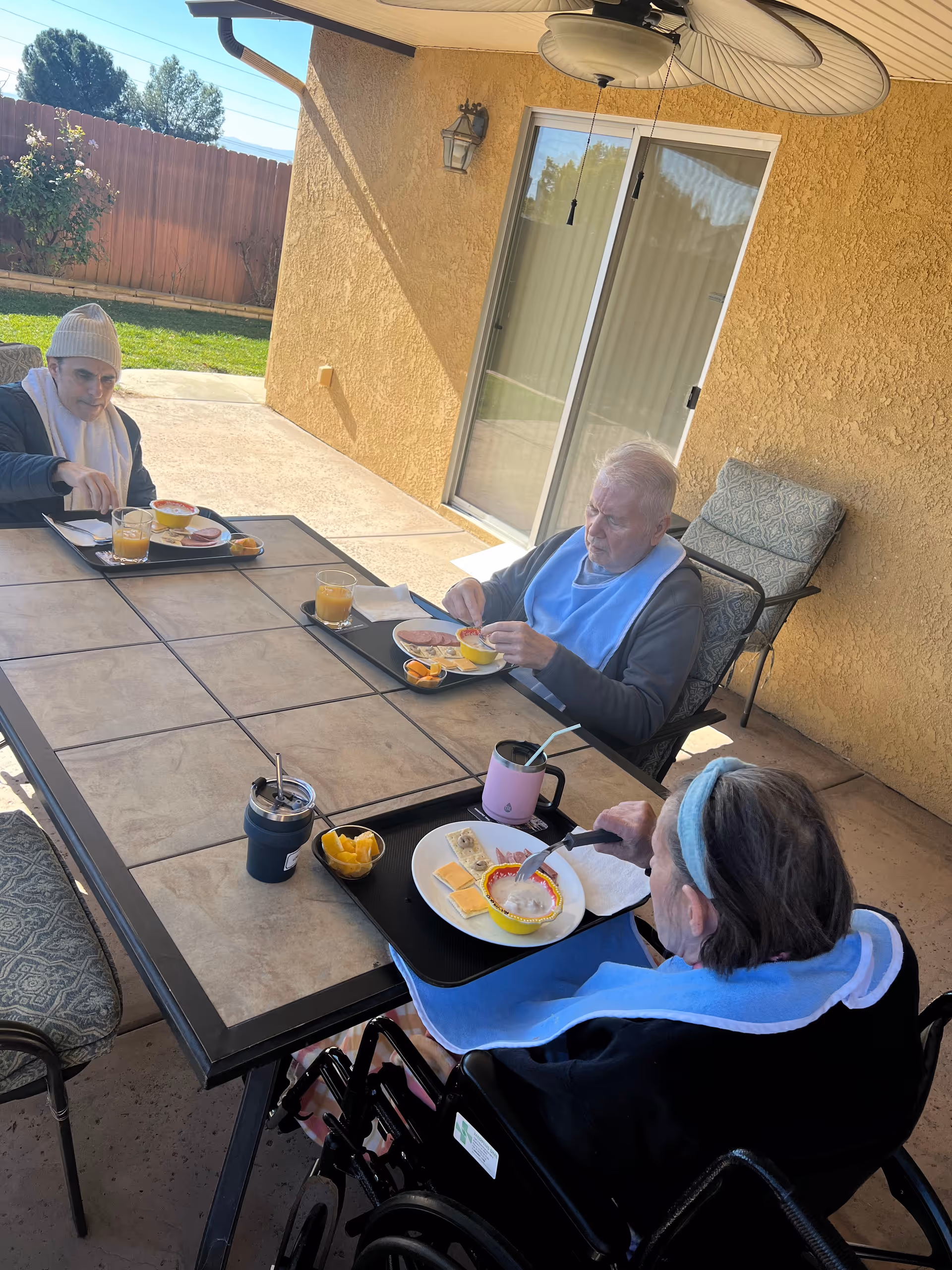 Three elderly individuals sitting around a rectangular outdoor table under a patio umbrella, eating a meal. Two are seated in chairs and one is in a wheelchair. The setting is a covered patio area with a sliding glass door and a yellow stucco wall in the background. The table has trays with plates of food and drinks.