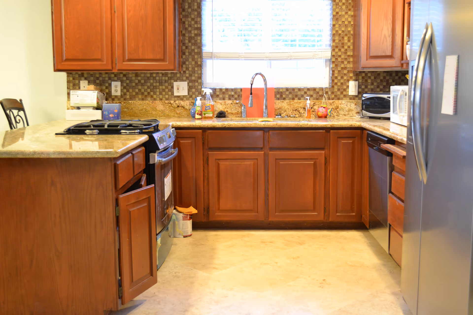 A kitchen with wooden cabinets, a granite countertop, a stainless steel refrigerator, a stove with an open cabinet door below it, a dishwasher, a microwave, and a window with blinds above the sink. Various cleaning supplies and kitchen items are placed on the countertop.