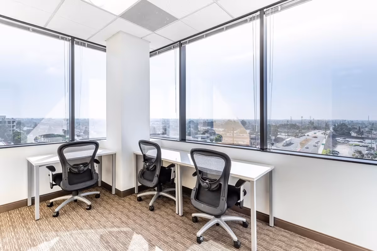 Corner office area with three desks, chairs, and large windows overlooking cityscape.