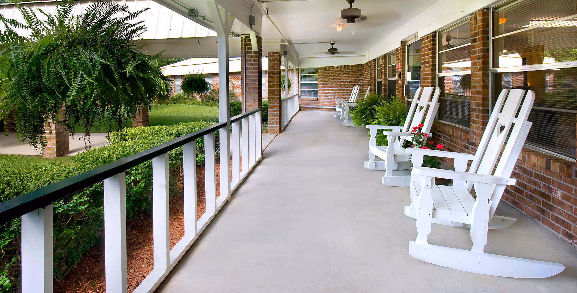 Covered outdoor veranda with white rocking chairs along a brick building.