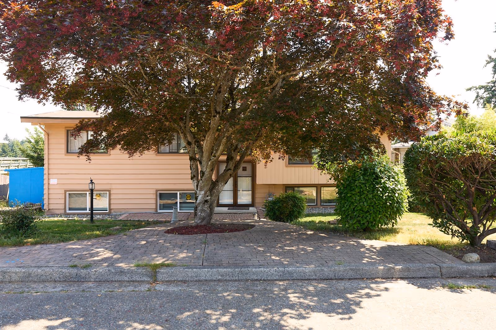 Front view of a beige, two-story senior care home with a large tree and shrubs.