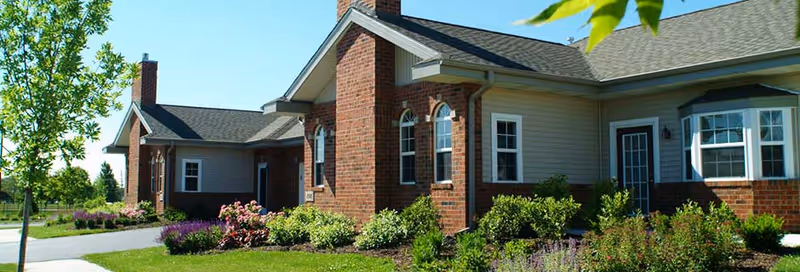 Front exterior of a single-story brick-and-siding building with landscaped shrubs and flowers under a clear blue sky.
