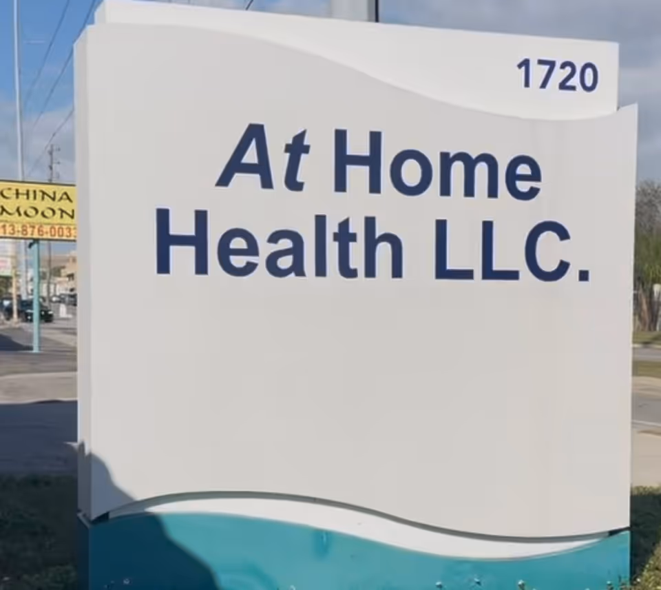 A large white sign reads At Home Health LLC with blue text beside a street.