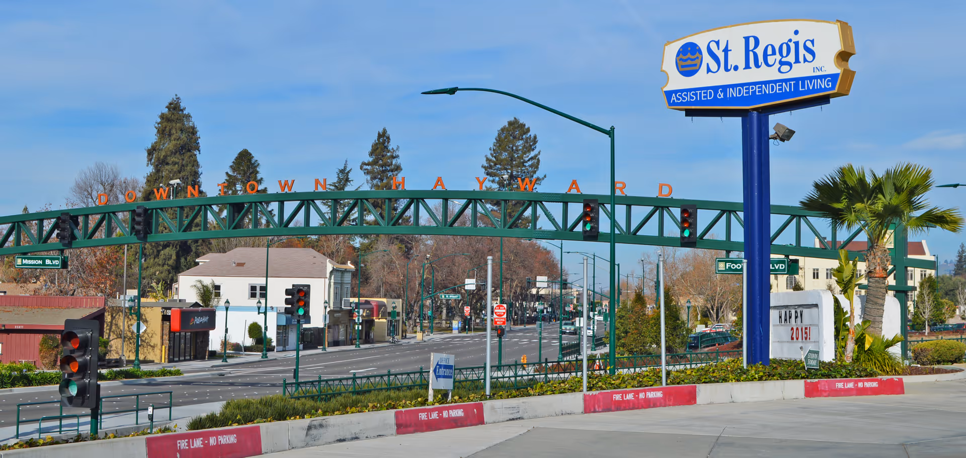 Street view of a downtown intersection featuring a large 'St. Regis Assisted & Independent Living' sign and a green 'Downtown Hayward' arch.