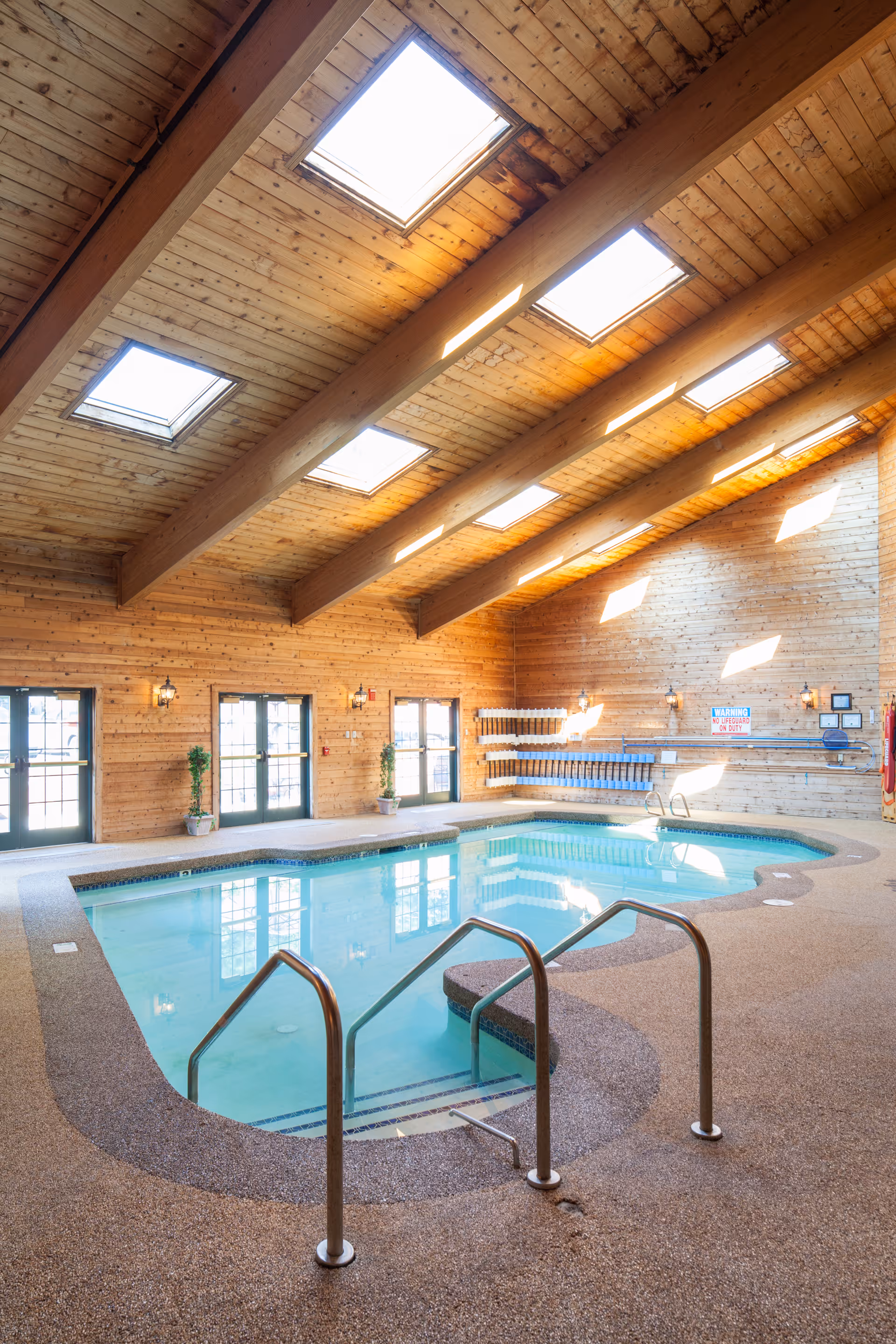 Indoor swimming pool with wooden walls and ceiling featuring skylights. The pool area has metal handrails and steps leading into the water. Several doors and potted plants are visible along the wall, with natural light streaming in.