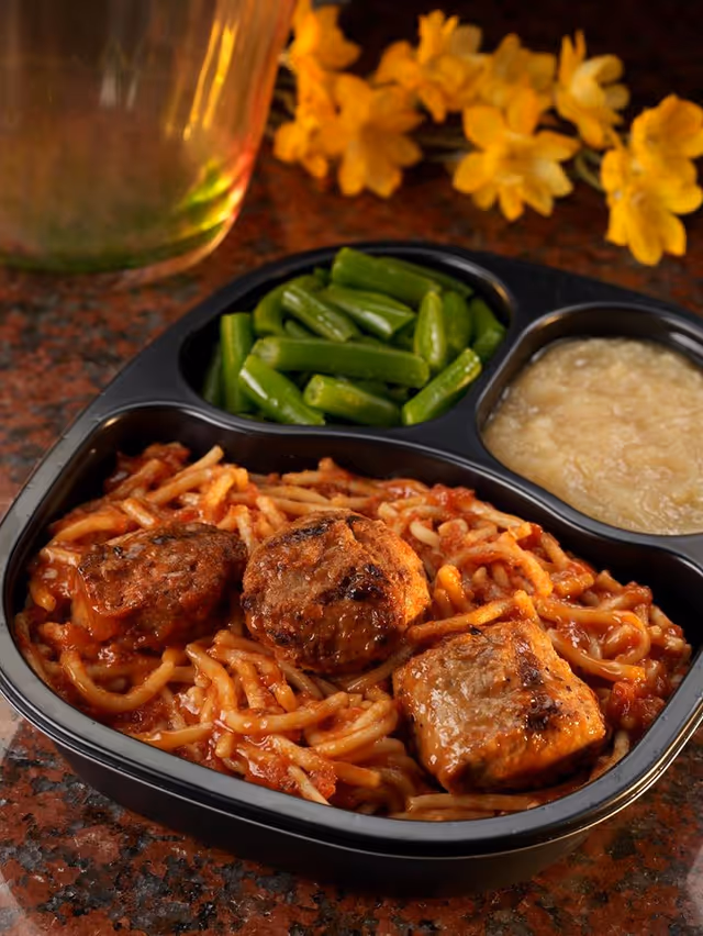 Close-up of a divided meal tray with noodles, meat, green beans, and sauce.
