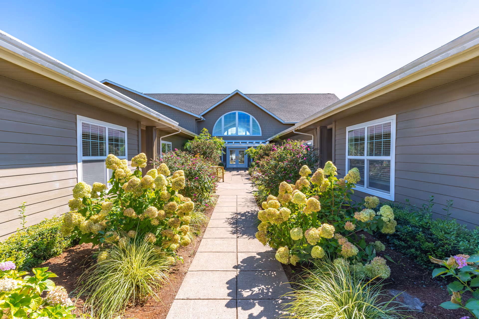 A paved walkway lined with flowering bushes and plants leads to the entrance of a single-story building with large windows and a peaked roof under a clear blue sky.
