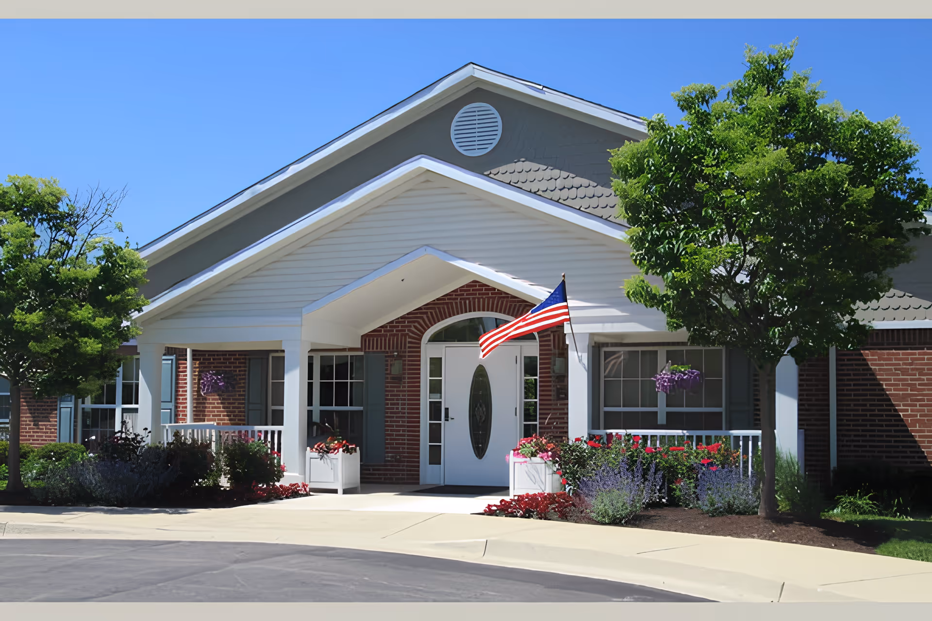 Front entrance of a brick senior living facility with a covered porch, American flag, and landscaped flower beds.