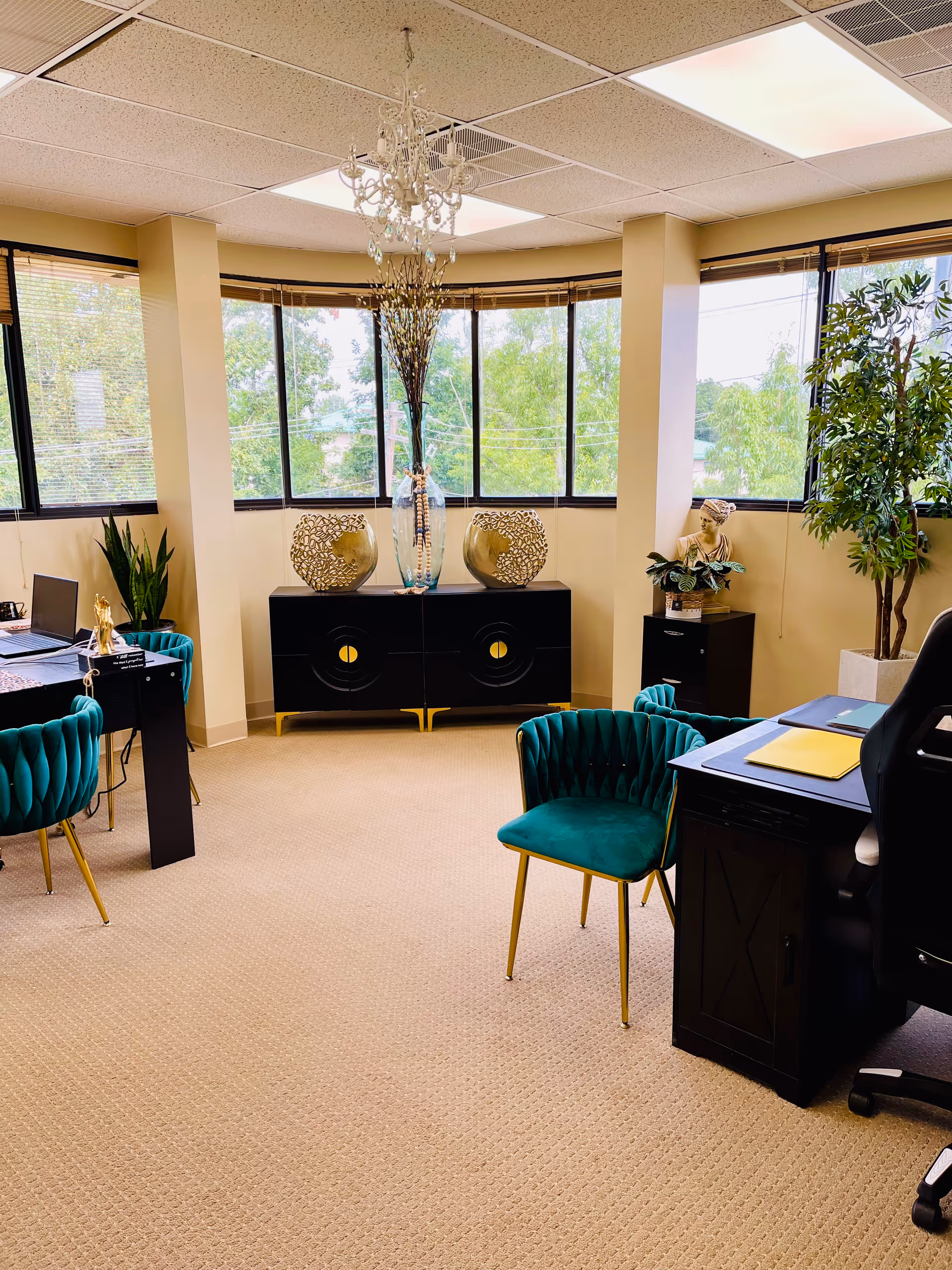 Curved-windowed lounge with teal chairs, a desk, chandelier, and decorative vases.