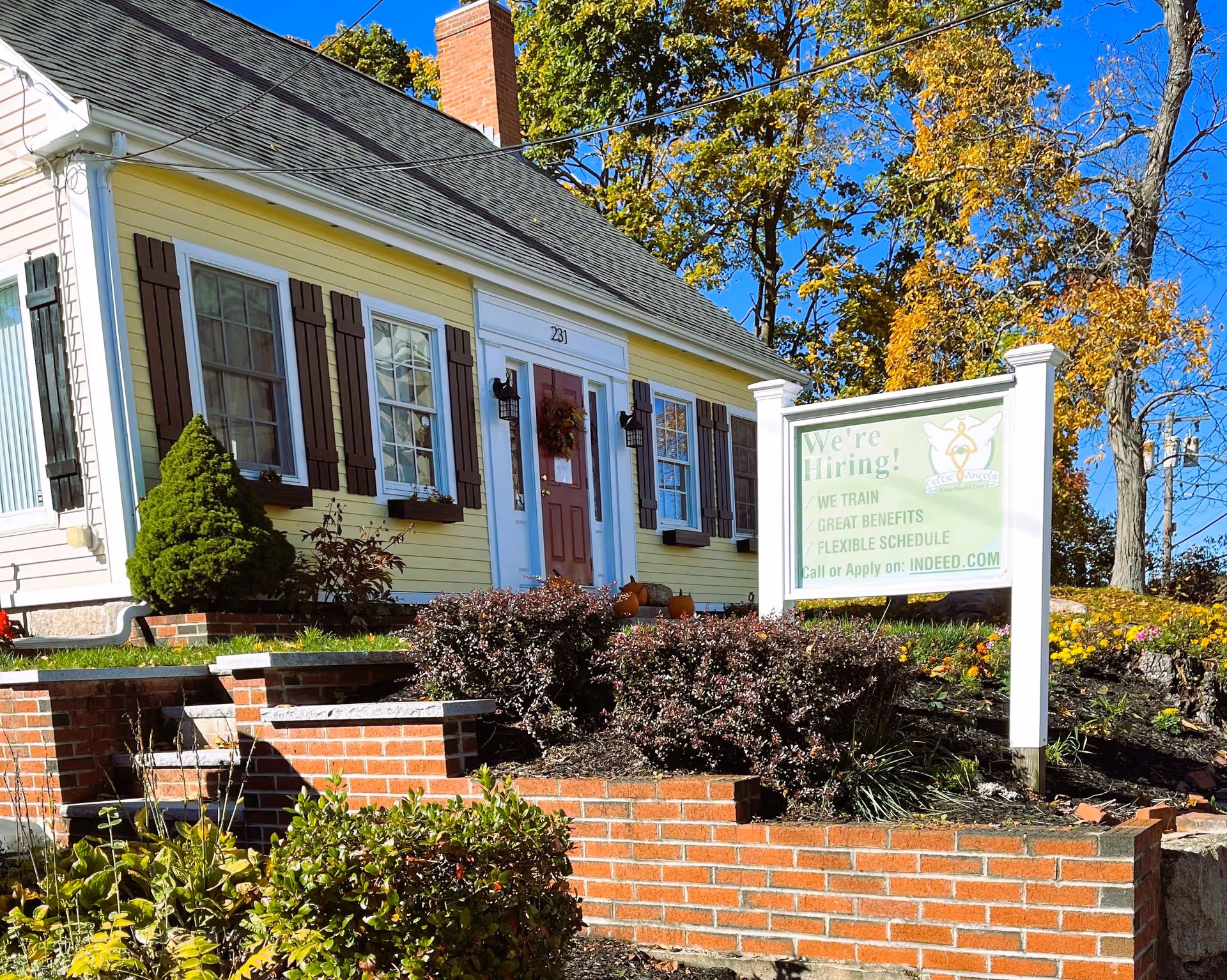 A yellow, house-style senior living building front with a hiring sign and landscaping.