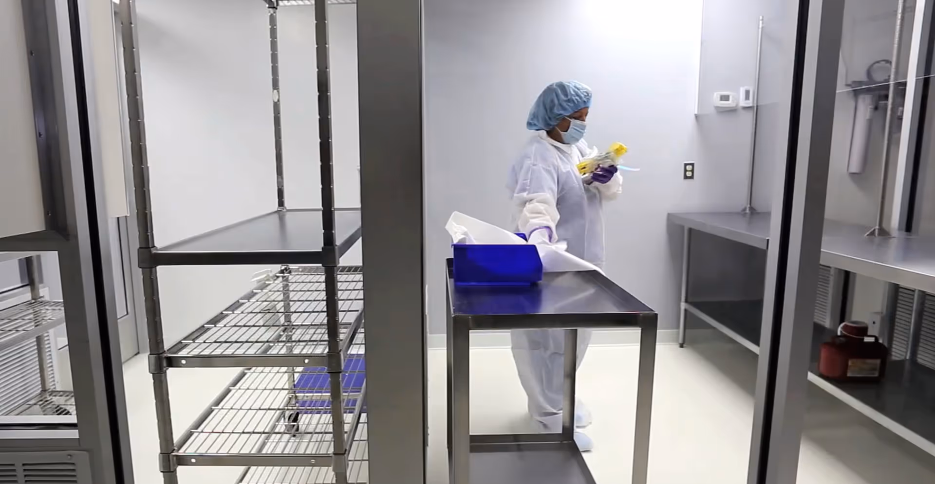 A worker in PPE inspects items in a sterile stainless steel facility room.