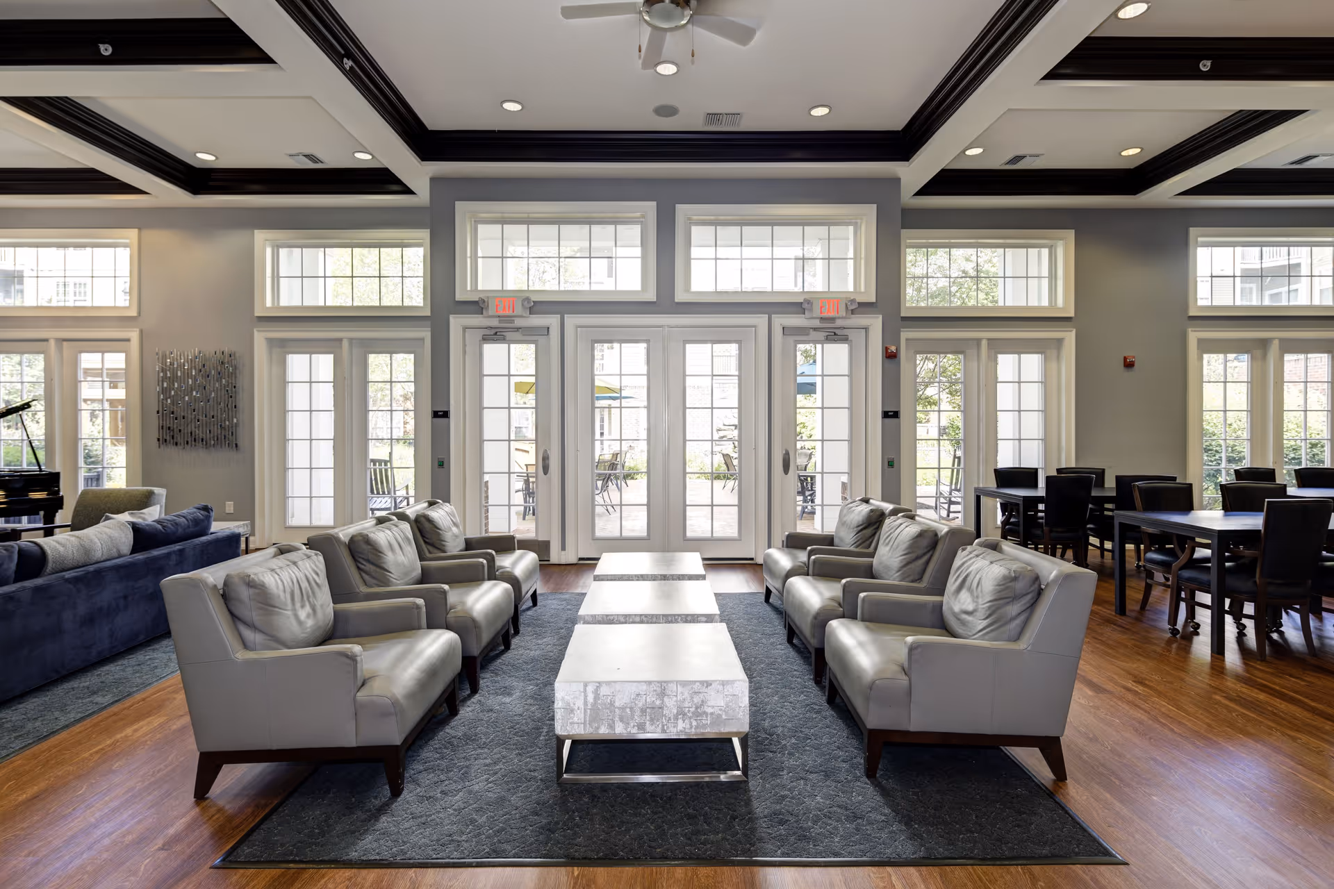 A spacious and well-lit common area with multiple gray armchairs arranged around three rectangular coffee tables on a dark rug. Large windows and glass doors let in natural light and provide a view of an outdoor patio with tables and chairs. To the right, there are several dining tables with black chairs. The ceiling features dark wooden beams contrasting with a white background.