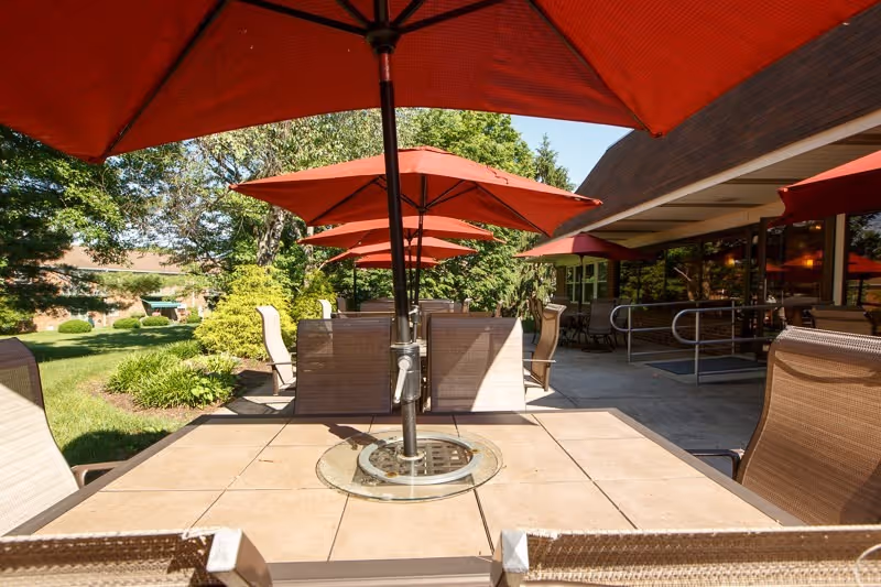 Outdoor patio area with multiple tables and chairs under large red umbrellas, surrounded by greenery and trees, adjacent to a building with large windows and a ramp.