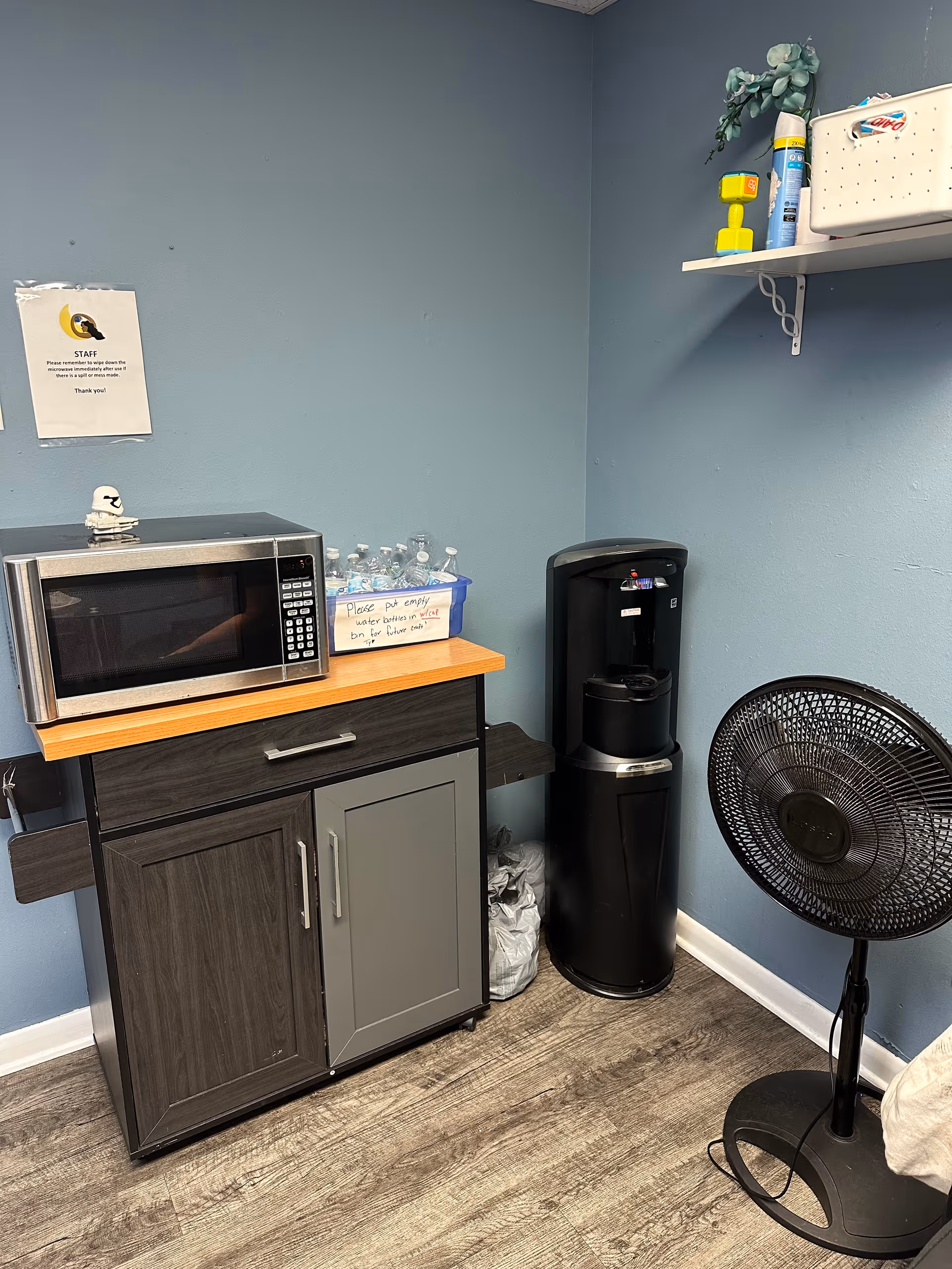 Kitchen corner with microwave on cabinet, water bottles, coffee maker, and a standing fan.