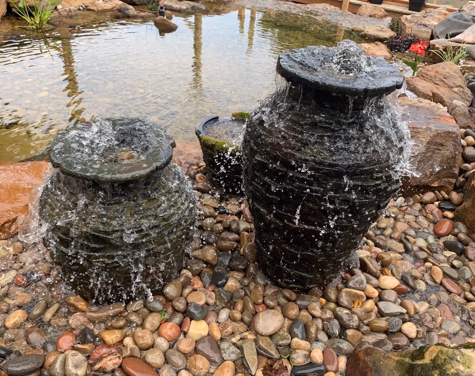 Two large stone urns with water flowing over their edges, placed on a bed of smooth multicolored pebbles next to a pond with clear water and rocks around the edge.