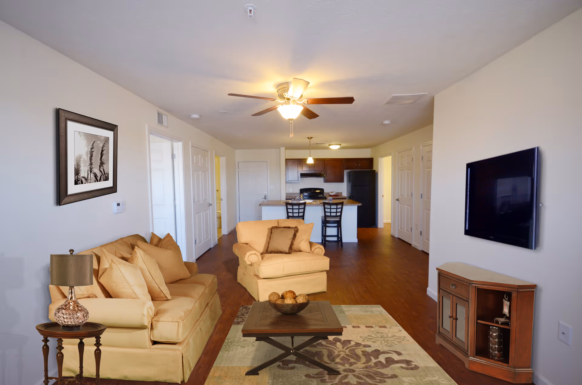 Open living room with beige sofa, coffee table, rug, wall mounted TV, and kitchen in back.