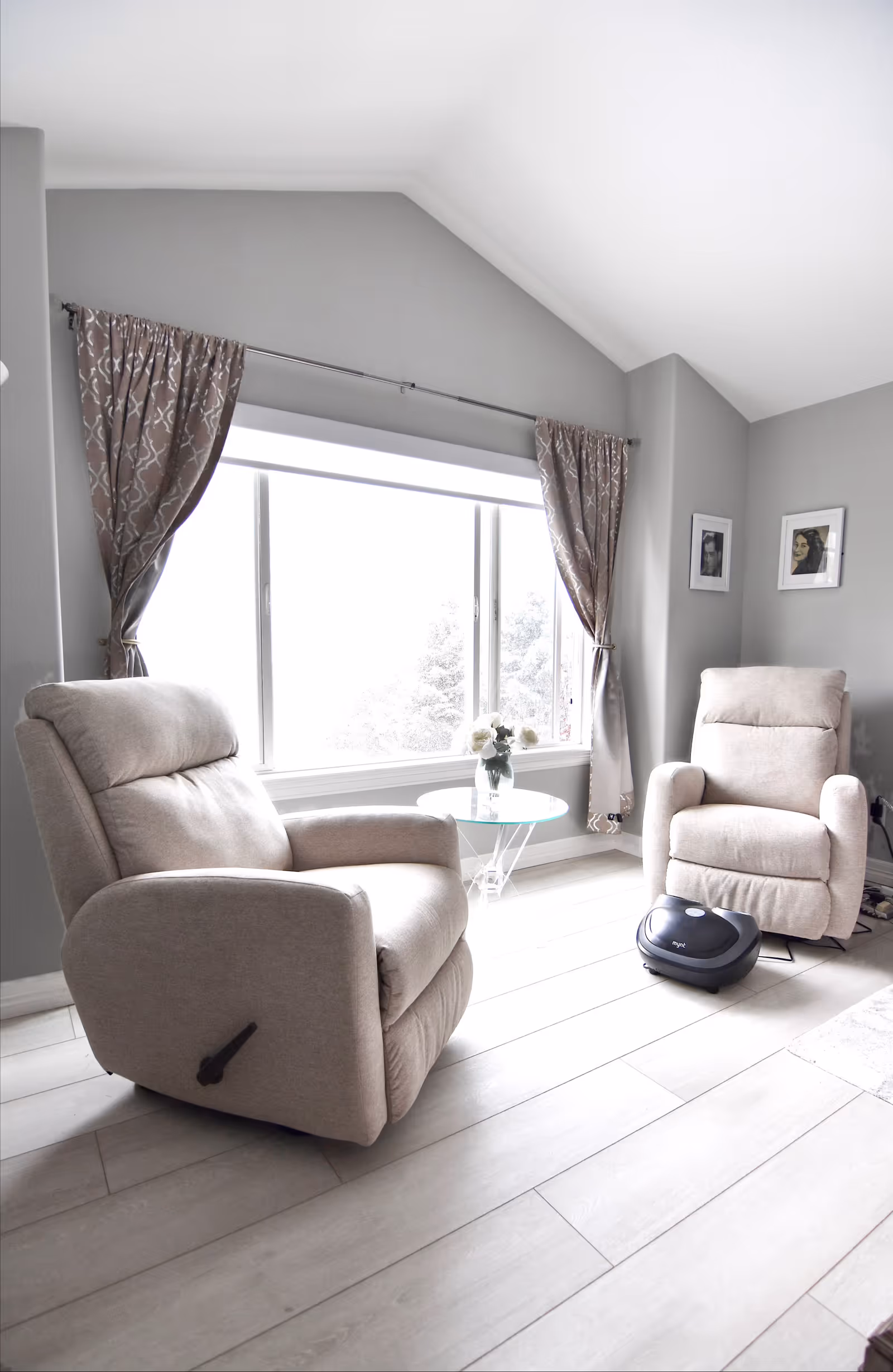 A bright living room with two beige recliner chairs facing each other near a large window with patterned curtains. A small round glass table with a vase of flowers is placed between the chairs. The room has light-colored wooden flooring and two framed pictures on the wall.