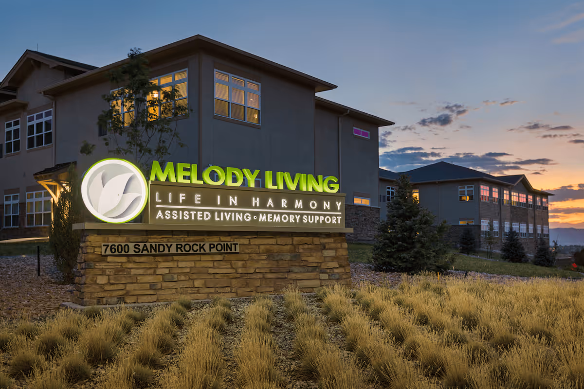Exterior view of Melody Living assisted living and memory support facility at sunset, showing the building and illuminated sign with address 7600 Sandy Rock Point.