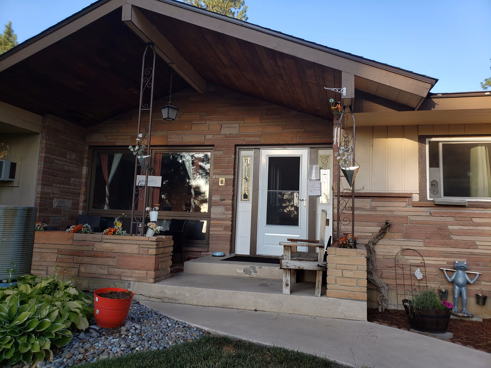 Front entrance of a single-story home with a covered porch, brick and wood exterior, a white door with glass panels, potted plants, a wooden bench, and decorative garden items including a metal frog statue.