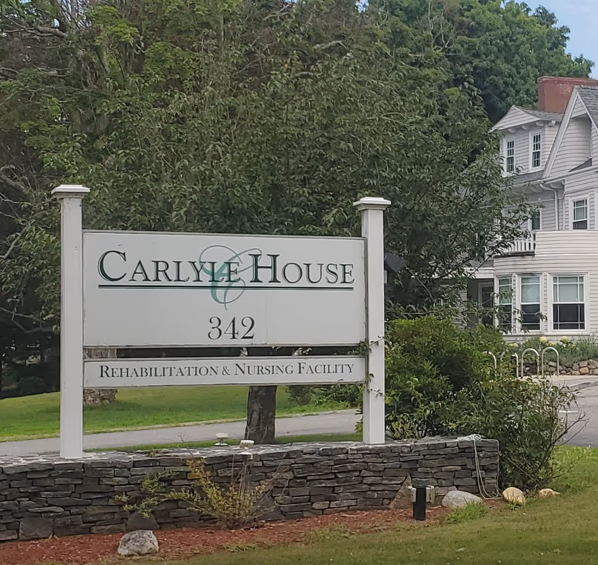 A white Carlyle House sign on a stone base with a building and trees behind.