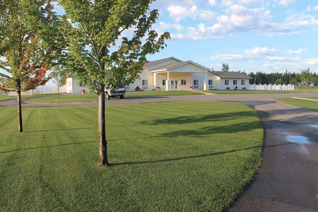 Single-story yellow memory care building with a curved driveway, manicured lawn and young trees under a blue sky.