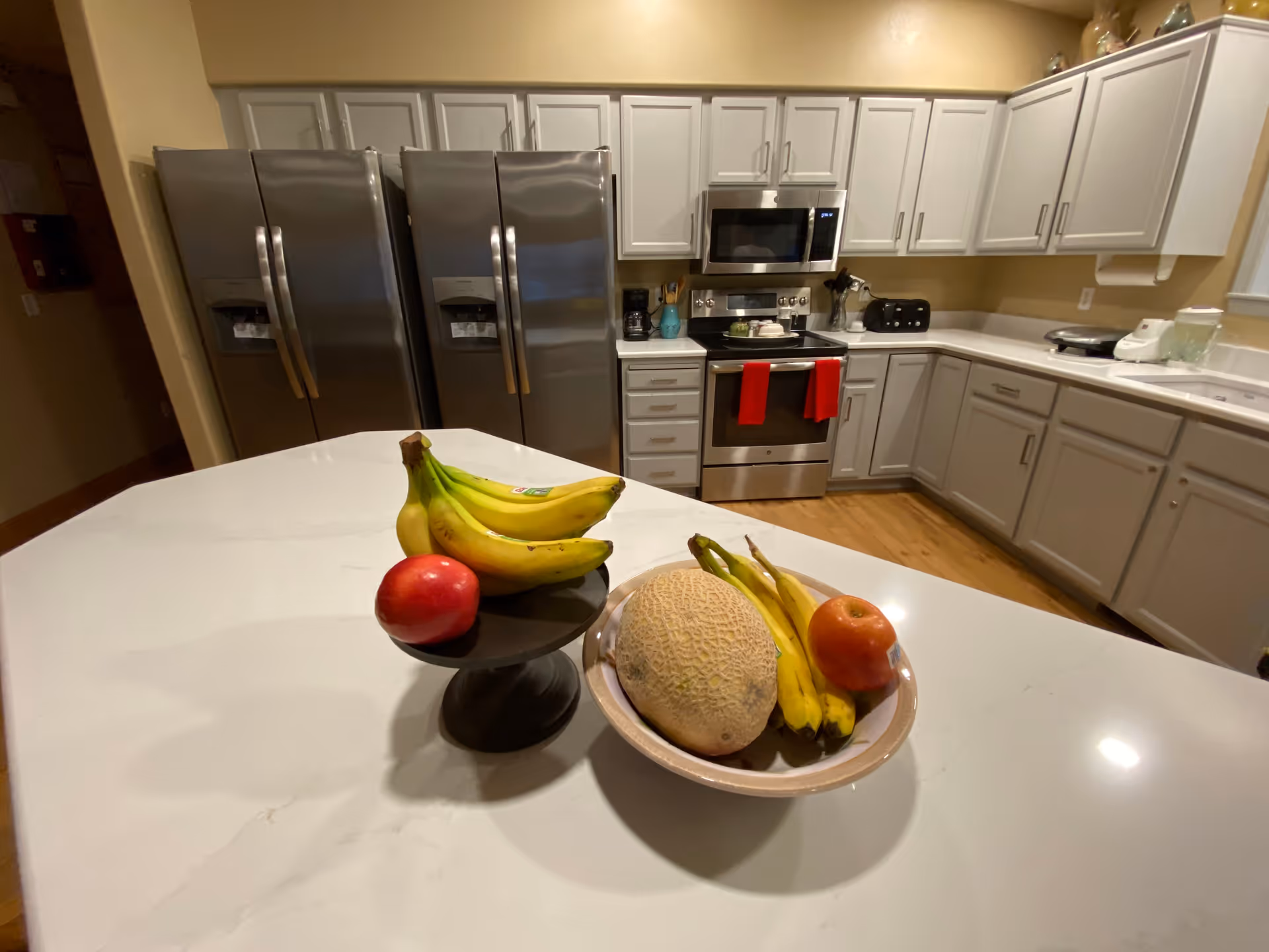 A modern kitchen with white cabinets and stainless steel appliances including two refrigerators, a microwave, and an oven. On the white countertop island in the foreground, there are two bowls of fruit containing bananas, a cantaloupe, and apples.