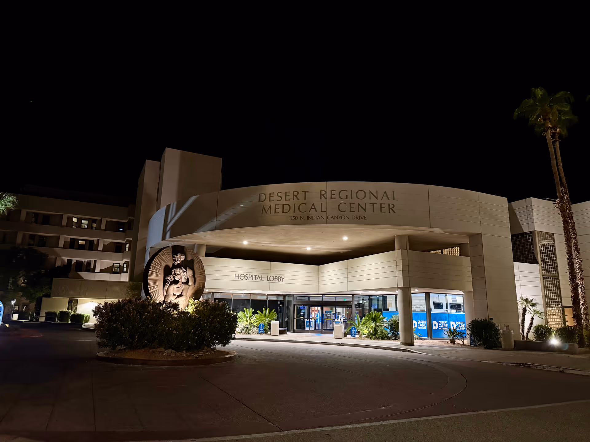 Night view of Desert Regional Medical Center's front entrance with sign and palm trees.