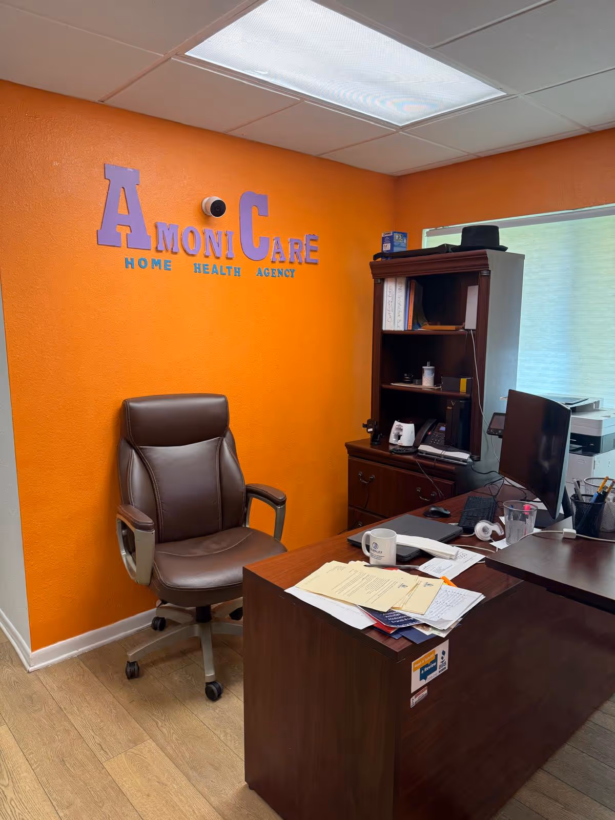 Office area with orange wall, Armoni Care signage, desk, chair, and computer equipment.