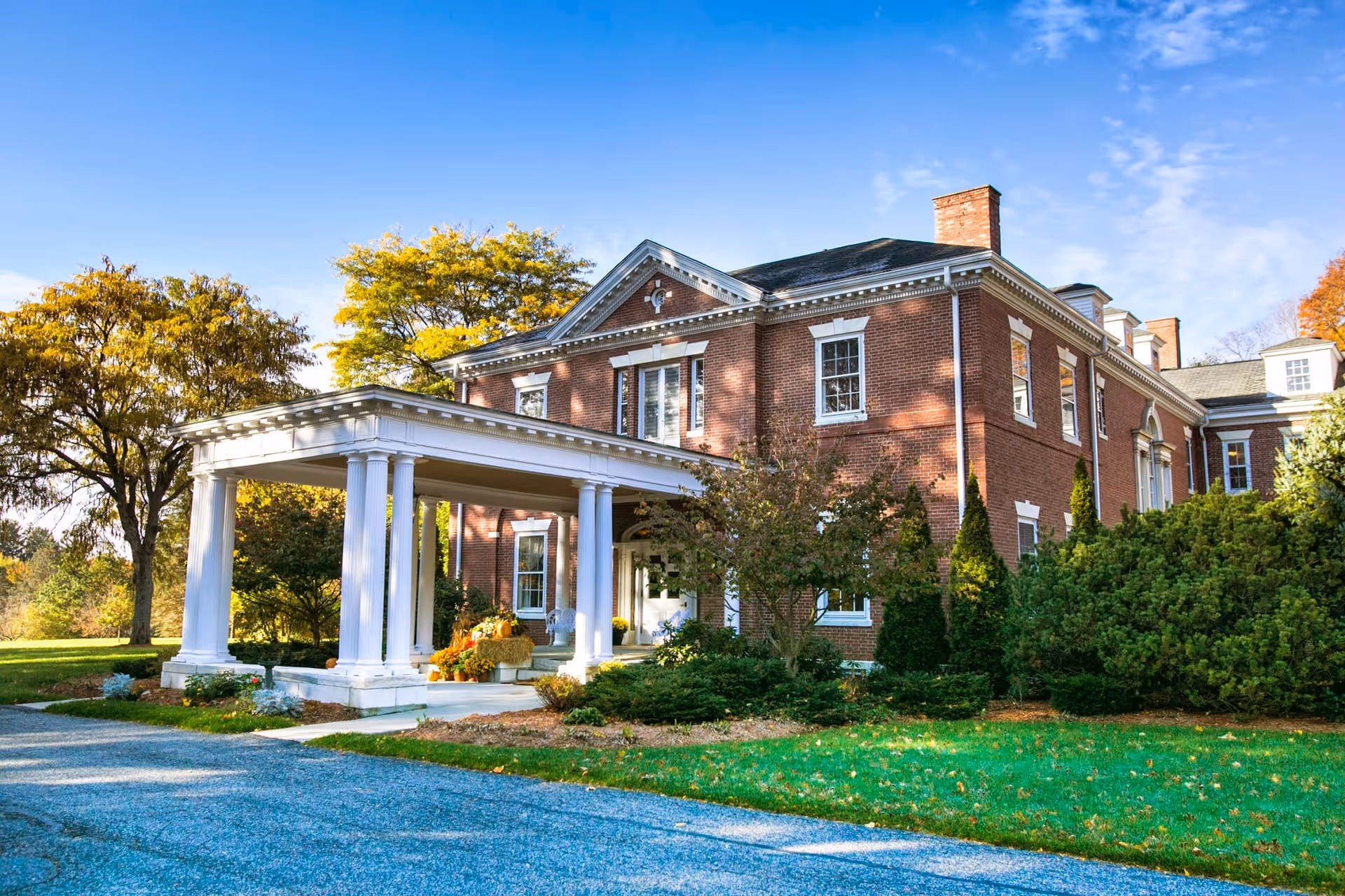 Two-story red brick building with a white columned portico entrance, surrounded by trees and a green lawn under a blue sky.