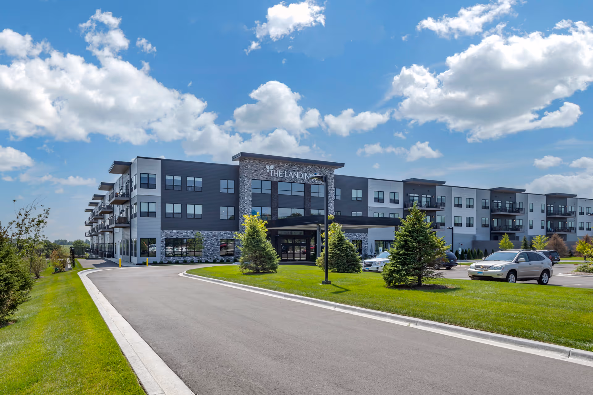 Exterior view of The Landings Senior Living facility on a sunny day with a clear blue sky and scattered clouds. The building is modern with three stories, featuring a combination of stone and siding on the facade. There is a driveway leading up to the entrance, surrounded by green grass and small trees. Several cars are parked in the parking lot to the right.