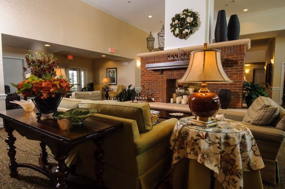 A cozy senior living facility common area with a brick fireplace, green sofas, a wooden table with a floral arrangement, and a side table with a lamp and floral tablecloth. The room is warmly lit with recessed ceiling lights and wall sconces, and decorated with plants and vases.