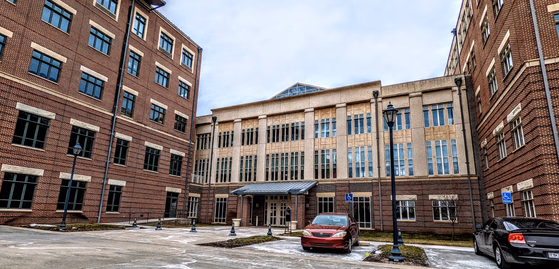 Brick senior living facility with central entrance, two wings, and parked cars under a blue sky.