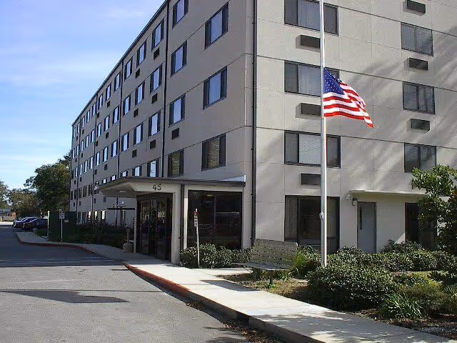 Exterior view of a multi-story senior living facility building with an entrance canopy marked with the number 45. There is an American flag on a flagpole in front of the building, along with a bench and landscaped bushes. The sky is clear and blue.