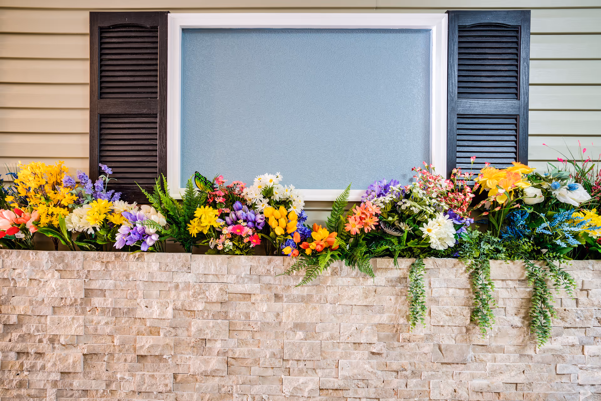 Colorful flower bed along a stone planter below a window with shutters on a building exterior.