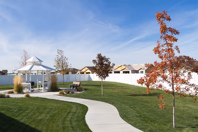 Sunny outdoor courtyard with white gazebo, curved walkway, benches, and trees near houses.