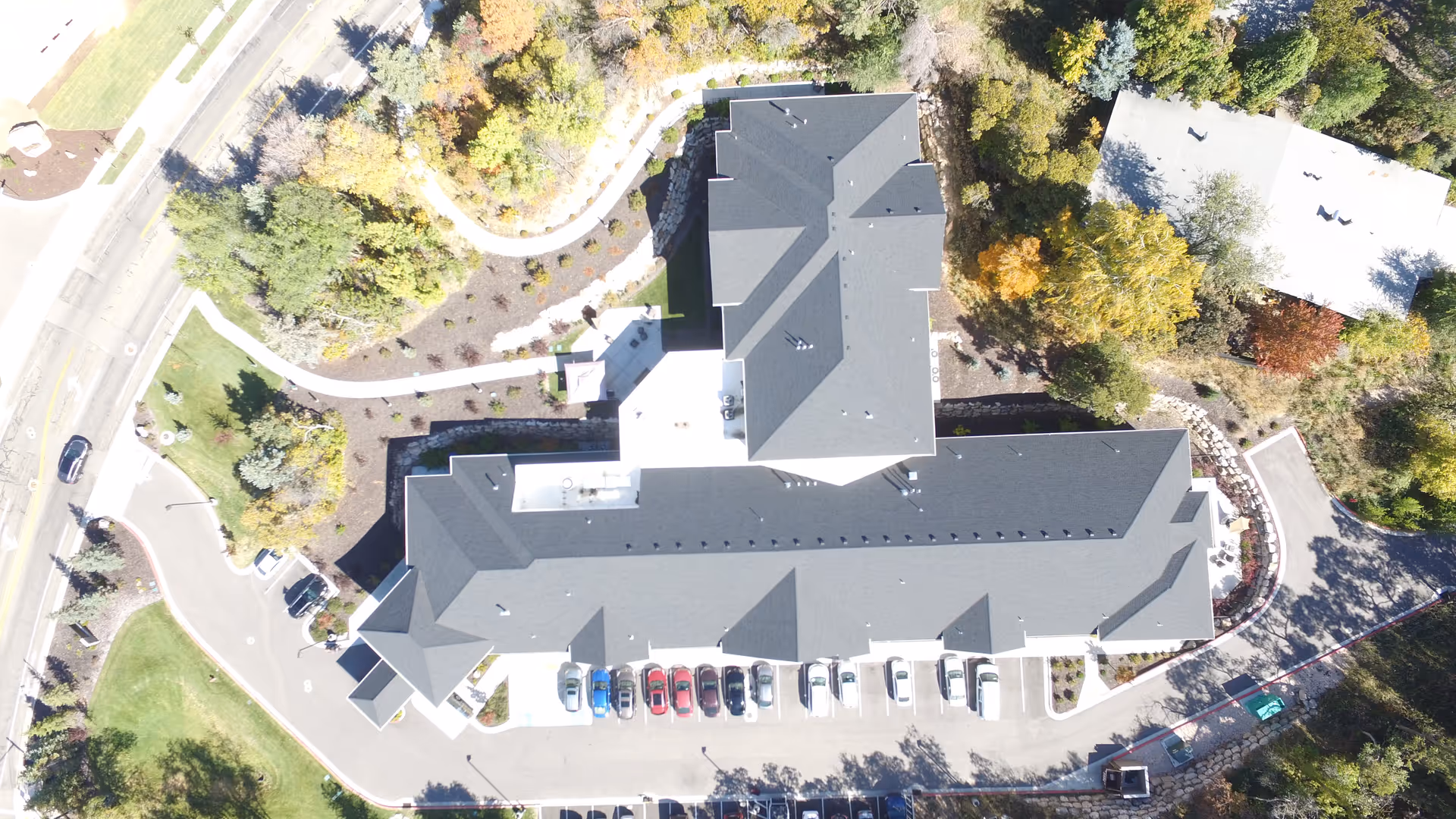 Aerial view of the Hidden Valley Assisted Living and Memory Care facility showing the building with a dark roof, surrounding parking lot with cars, landscaped greenery, trees, and adjacent roads.