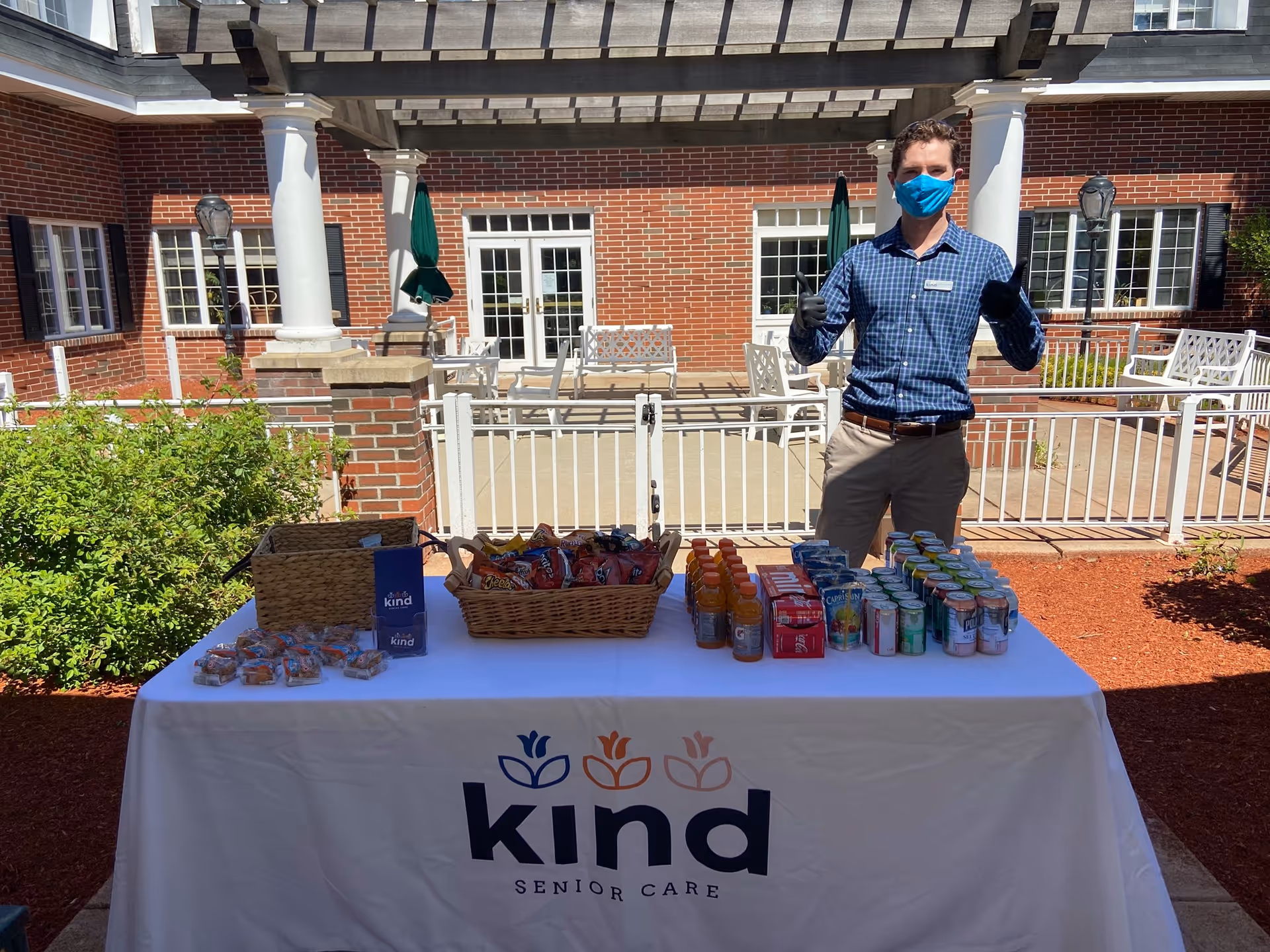 Staff member wearing a mask stands behind a snack table on an outdoor senior care courtyard.