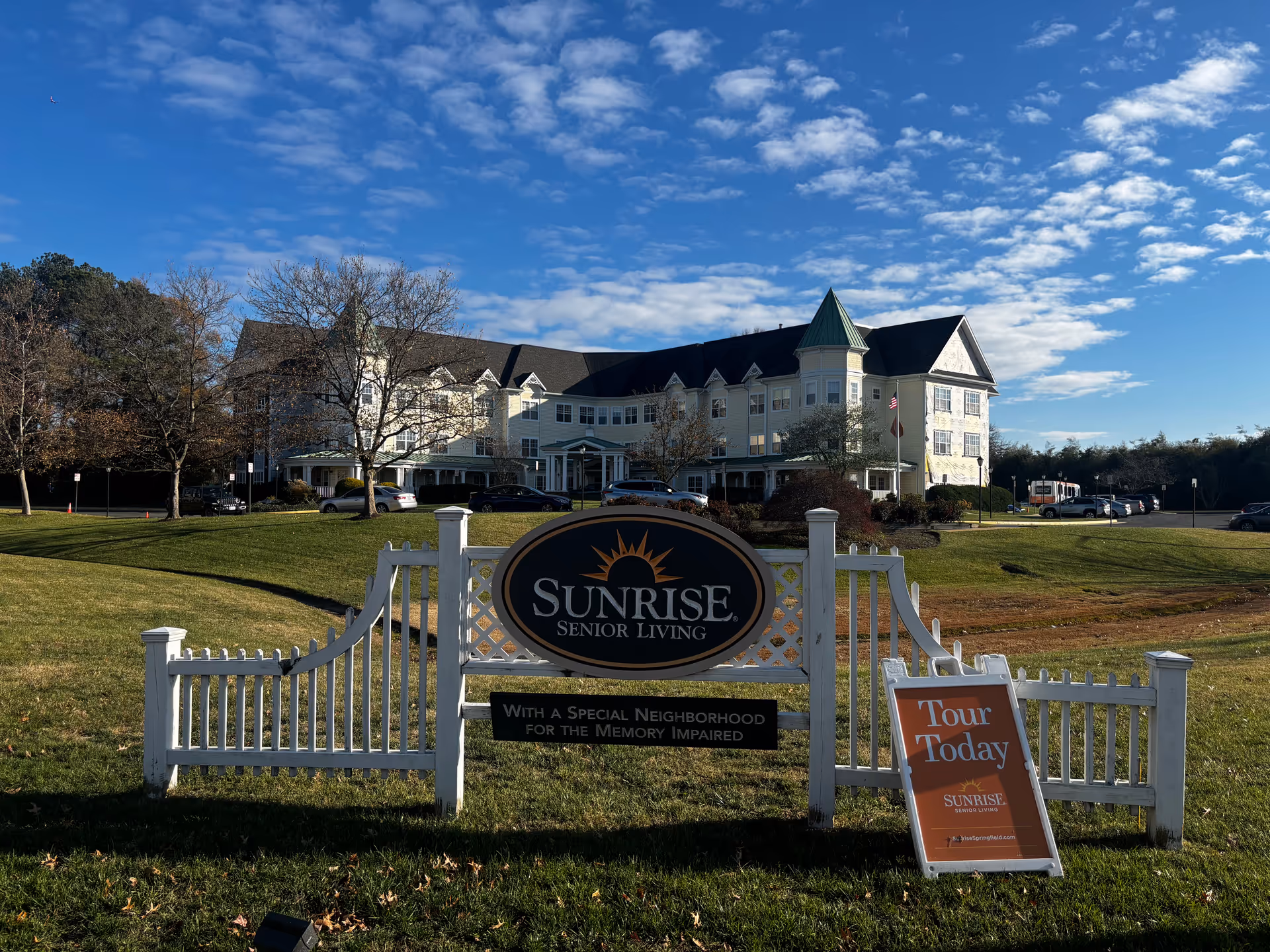 Front exterior of the Sunrise Senior Living building with a prominent entrance sign and a 'Tour Today' sandwich board on a grassy lawn.