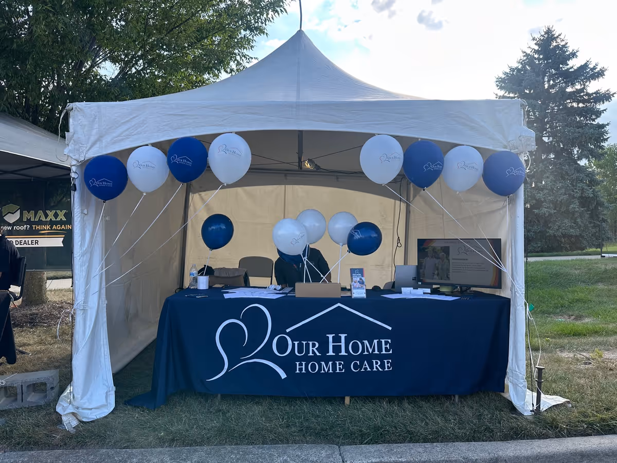 Outdoor tent booth with balloons and a table displaying Our Home Home Care branding.