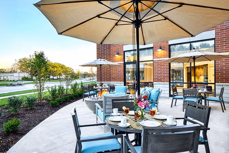 Outdoor patio area at Sheridan at Park Ridge with round tables set for dining, chairs with blue cushions, large beige umbrellas providing shade, a fire pit, and a brick building with large windows in the background. There are plants and trees along the edge of the patio.