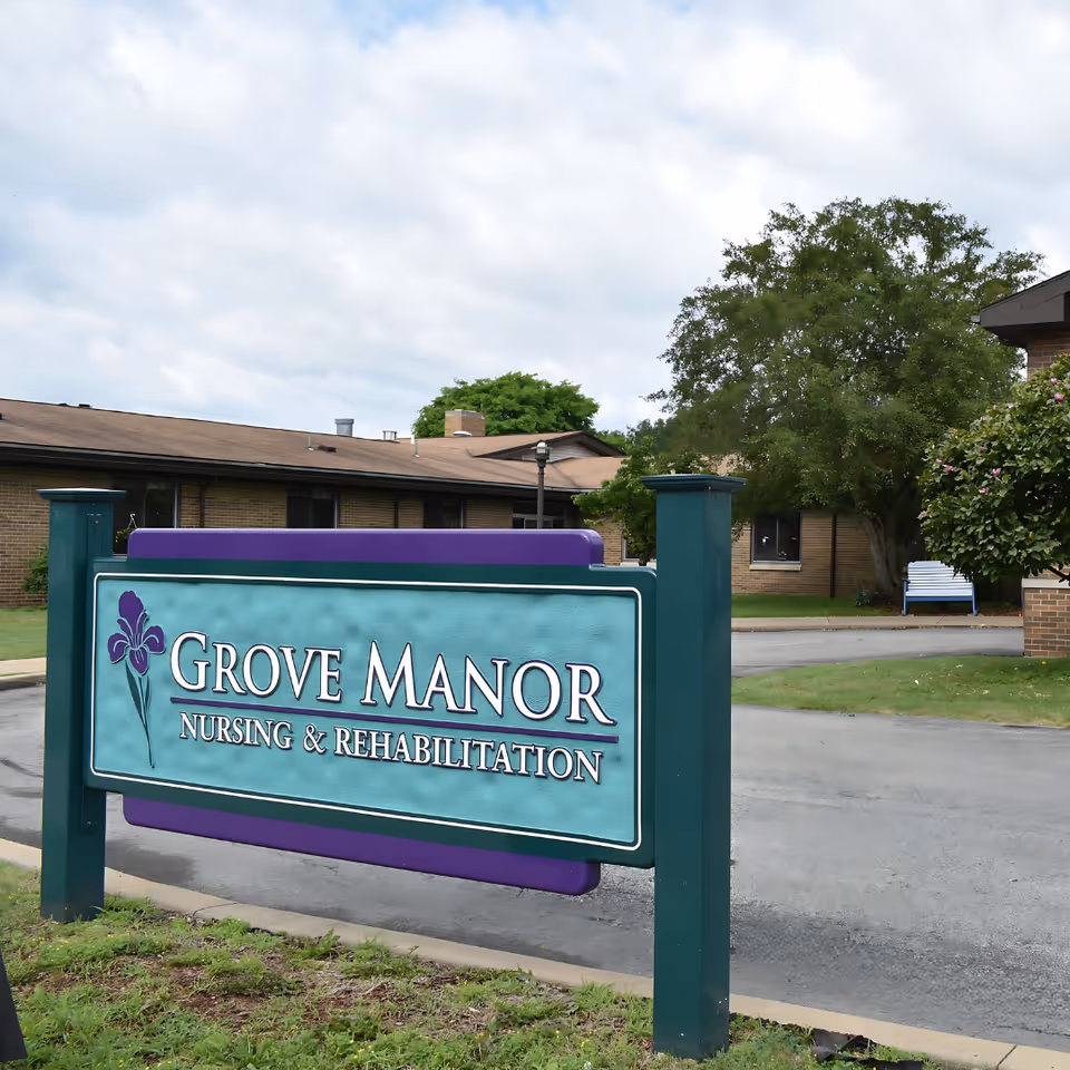 Outdoor view of Grove Manor Nursing & Rehabilitation facility sign with a building and trees in the background under a cloudy sky.