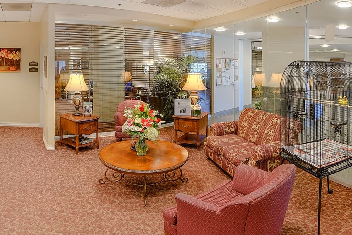 A furnished seating area with patterned sofa and chairs around a round wooden coffee table topped with a floral arrangement, lamps and side tables, and a large birdcage in a memory care facility lobby.