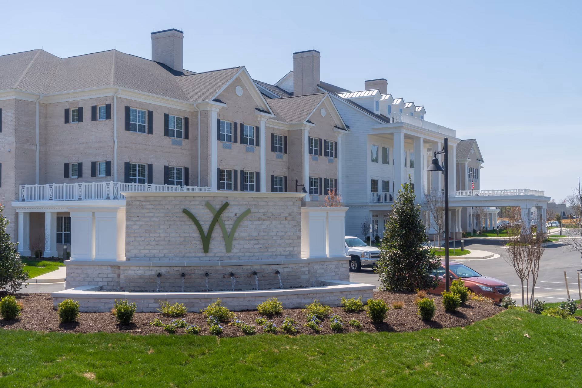 Exterior view of The Westmont at Short Pump senior living facility showing a large multi-story building with beige brick and white siding, multiple windows with black shutters, a landscaped area with green grass and shrubs, a stone sign with a green 'W' logo, and a driveway with parked cars under a clear blue sky.