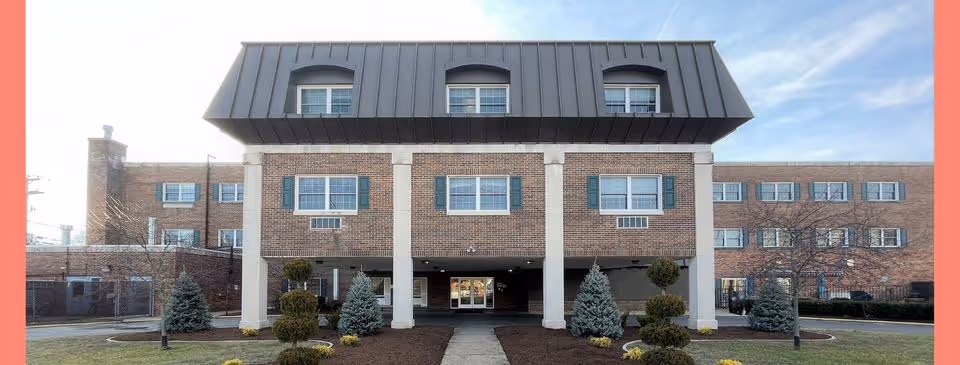 Front exterior view of a three-story brick building with a covered entrance supported by four white columns. The building has multiple windows with green shutters and a dark metal mansard roof. There are landscaped bushes and small trees in front of the building, and a concrete walkway leads to the entrance.