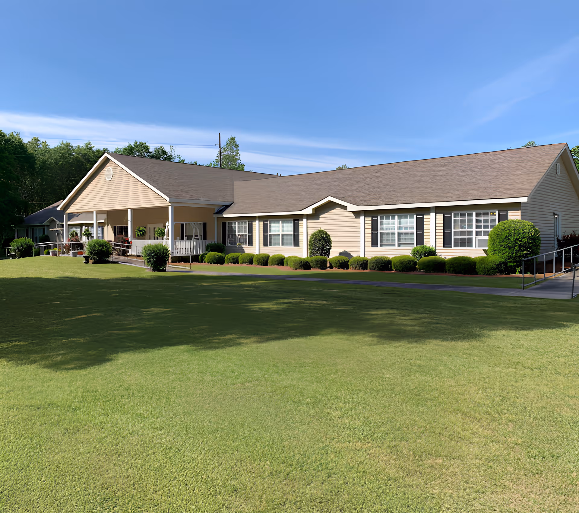 Single-story beige building with a covered porch and trimmed shrubs set behind a large green lawn under a clear blue sky.