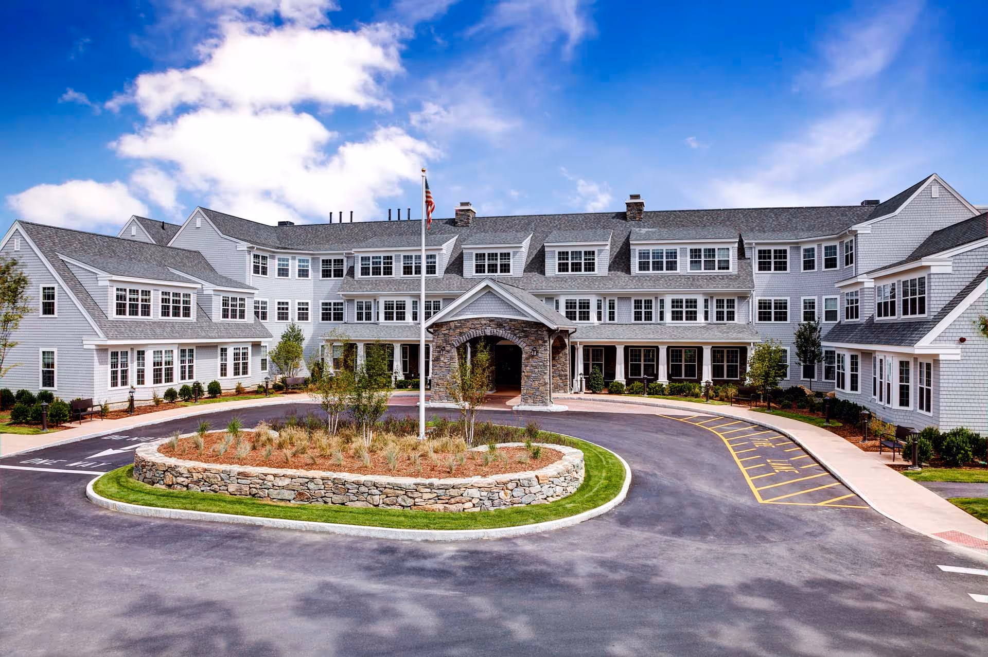 Front exterior of a multi-story senior living facility with a circular driveway, landscaped island, and flagpole under a blue sky.